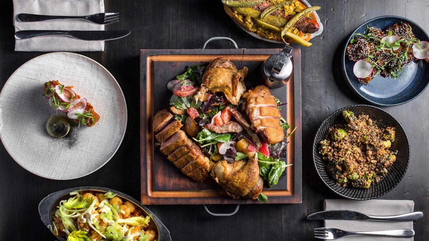 A spread of food on a table centered on a wooden board with a rotisserie duck