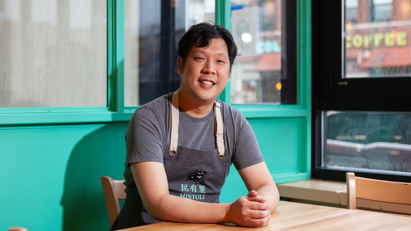 A seated Rich Wang smiles in an apron in front of a seafoam wall and windows