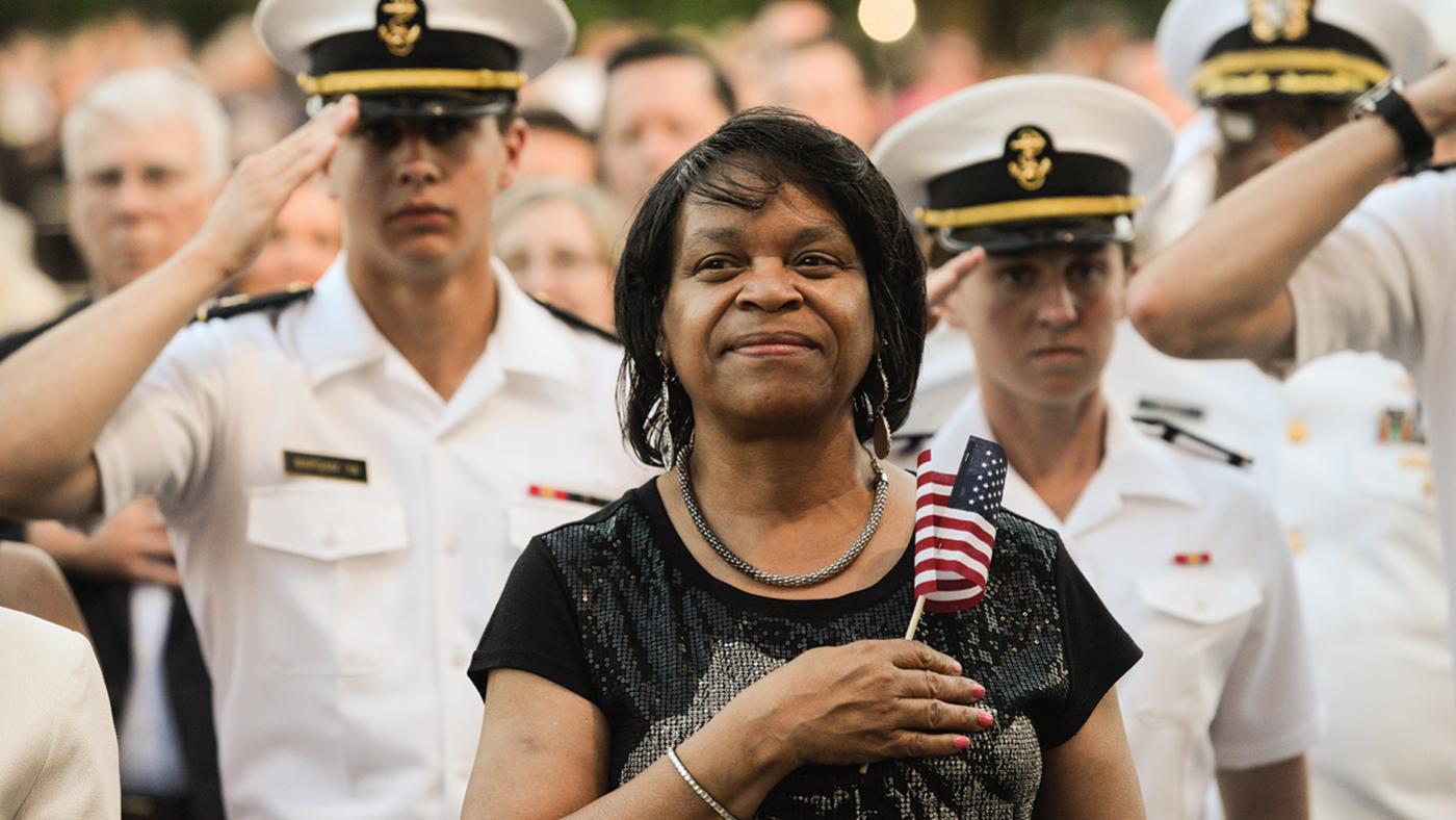 A woman stands holding an American flag over her heart in front of military people in uniform