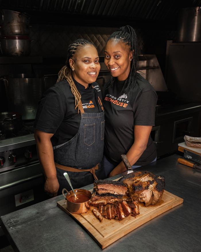 Dominique Leach standing with her wife, Tanisha Griffin, in front of a board of barbecue. Image: Provided