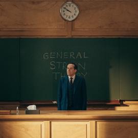 Professor T stands behind a desk in front of a chalkboard in a lecture hall