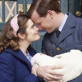 Helen and James smile and hold their heads to each other as James holds their newborn